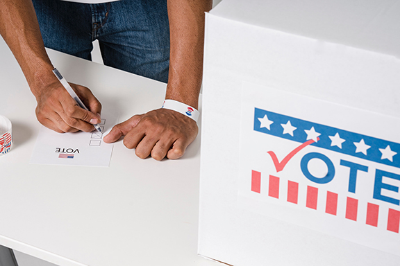 Person checking boxes on a ballot beside a dropbox.